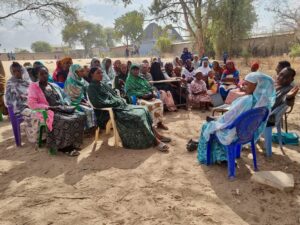 A group of women and children are seated in a circle on plastic chairs outdoors in a sandy area, engaged in a discussion. Trees and a few buildings are visible in the background.