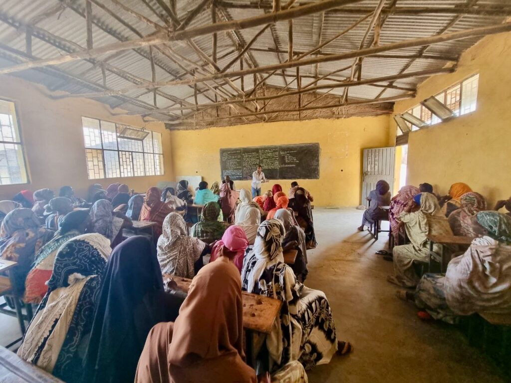 A classroom filled with women wearing colorful headscarves, seated at desks and facing a chalkboard, while a teacher stands at the front of the room. The classroom has yellow walls and a corrugated metal roof.