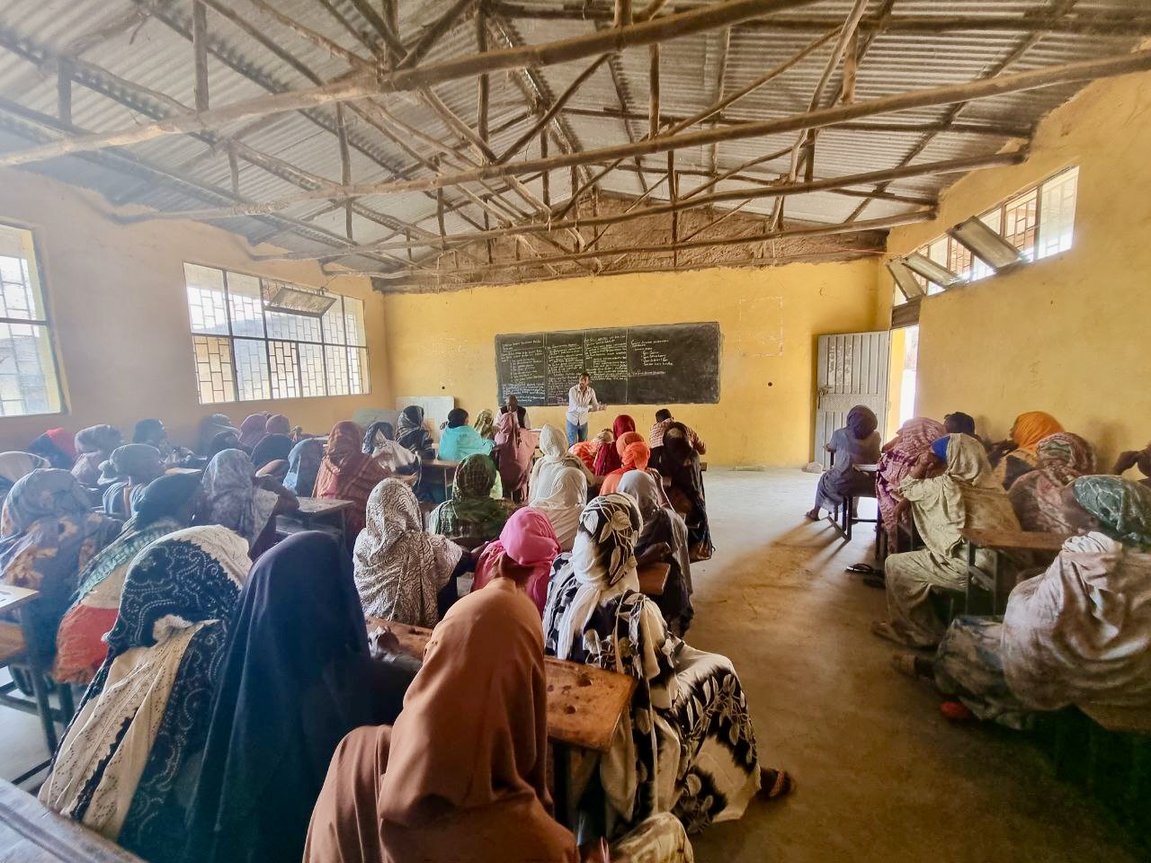 A classroom filled with women wearing colorful headscarves, seated at desks and facing a chalkboard, while a teacher stands at the front of the room. The classroom has yellow walls and a corrugated metal roof.