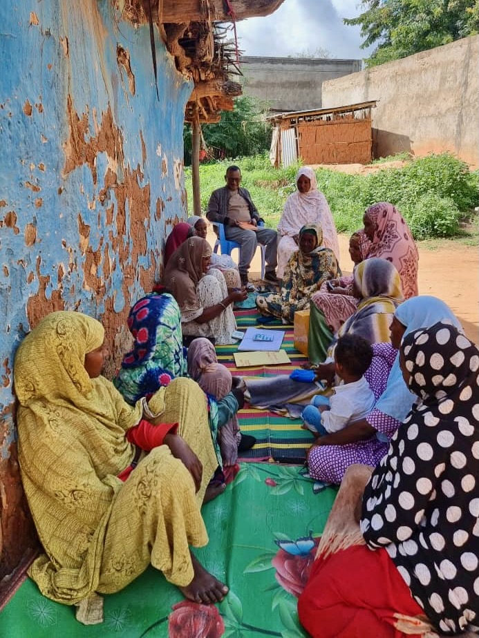 A group of women and children sit on mats in a circle outdoors near a worn blue wall, with a man seated on a chair at the far end, engaged in a discussion or meeting in a rural setting.