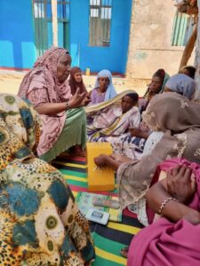 A group of women sit together on colorful mats outside, engaged in a discussion. One woman speaks animatedly while others listen attentively, holding notebooks and papers. A bright blue building is in the background.