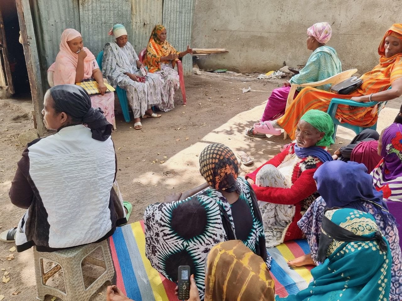 A group of women in colorful clothing sit together outdoors on plastic chairs and mats, engaging in conversation near a corrugated metal wall. Sunlight filters onto the ground, and some appear to be listening attentively.