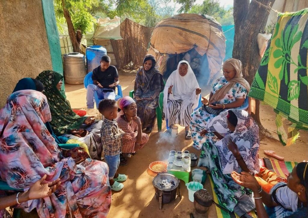 A group of women and children sit outdoors in a circle around a coffee ceremony setup, with cups and a pot on a small stove. Some people are seated on mats, while others sit on chairs under the shade of trees.