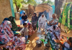 A group of women and children sit outdoors in a circle around a coffee ceremony setup, with cups and a pot on a small stove. Some people are seated on mats, while others sit on chairs under the shade of trees.