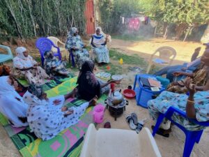 A group of women in colorful hijabs sit on mats and chairs around a small fire, drinking tea in a courtyard.