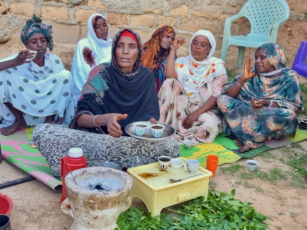 A group of women in colorful clothing sit on mats outdoors, gathered around a traditional coffee ceremony setup with cups, leaves, and a charcoal stove, against a backdrop of a stone wall and plastic chairs.